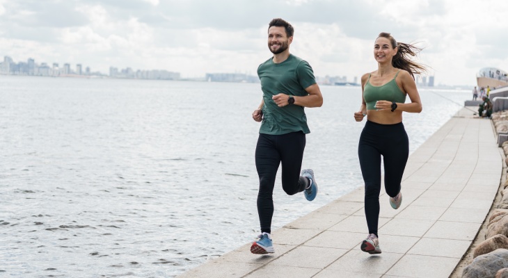 A couple running along a body of water with a city landscape in the background.